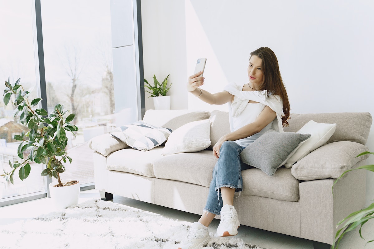 Positive-young-female-doing-selfie-on-smartphone-while-sitting-on-sofa